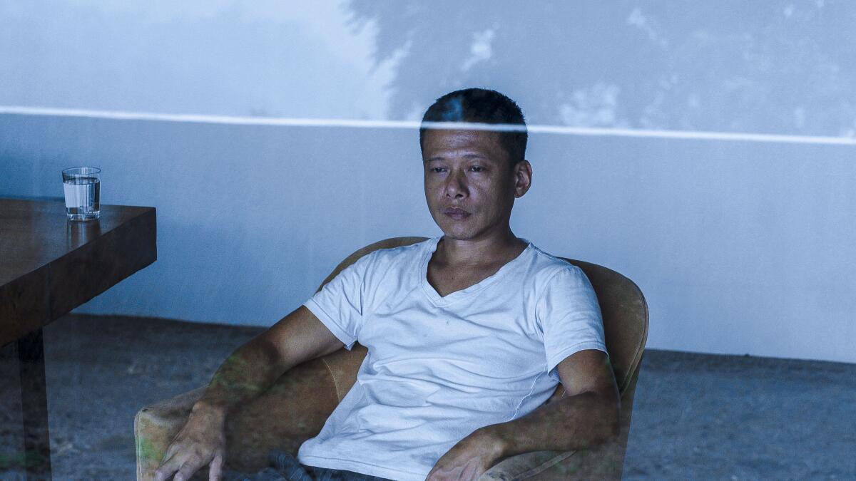 A man in a white T-shirt sits in a chair staring out a window.