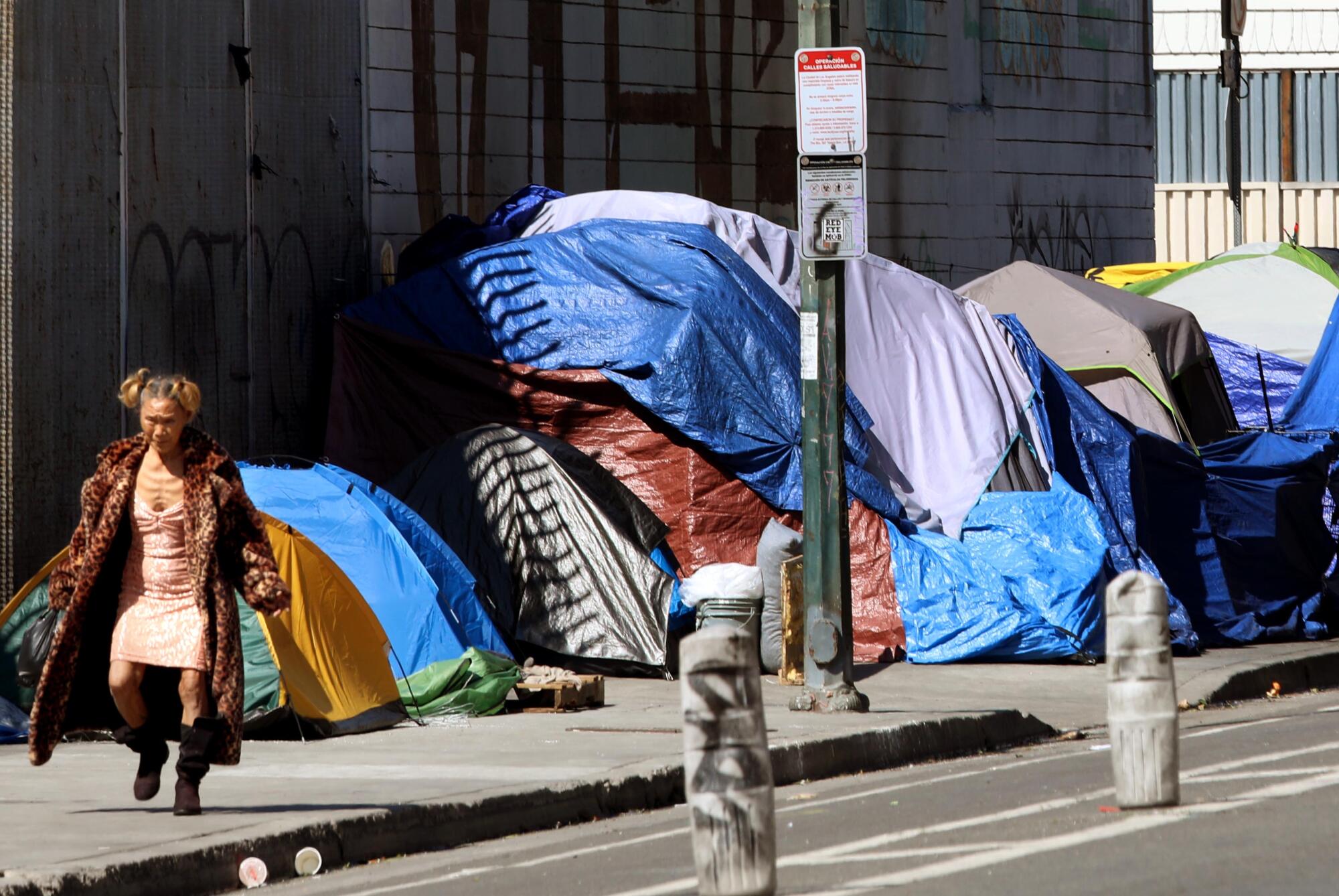 March 2025 photo of a homeless woman walking past a row of tents in Skid Row in downtown Los Angeles.