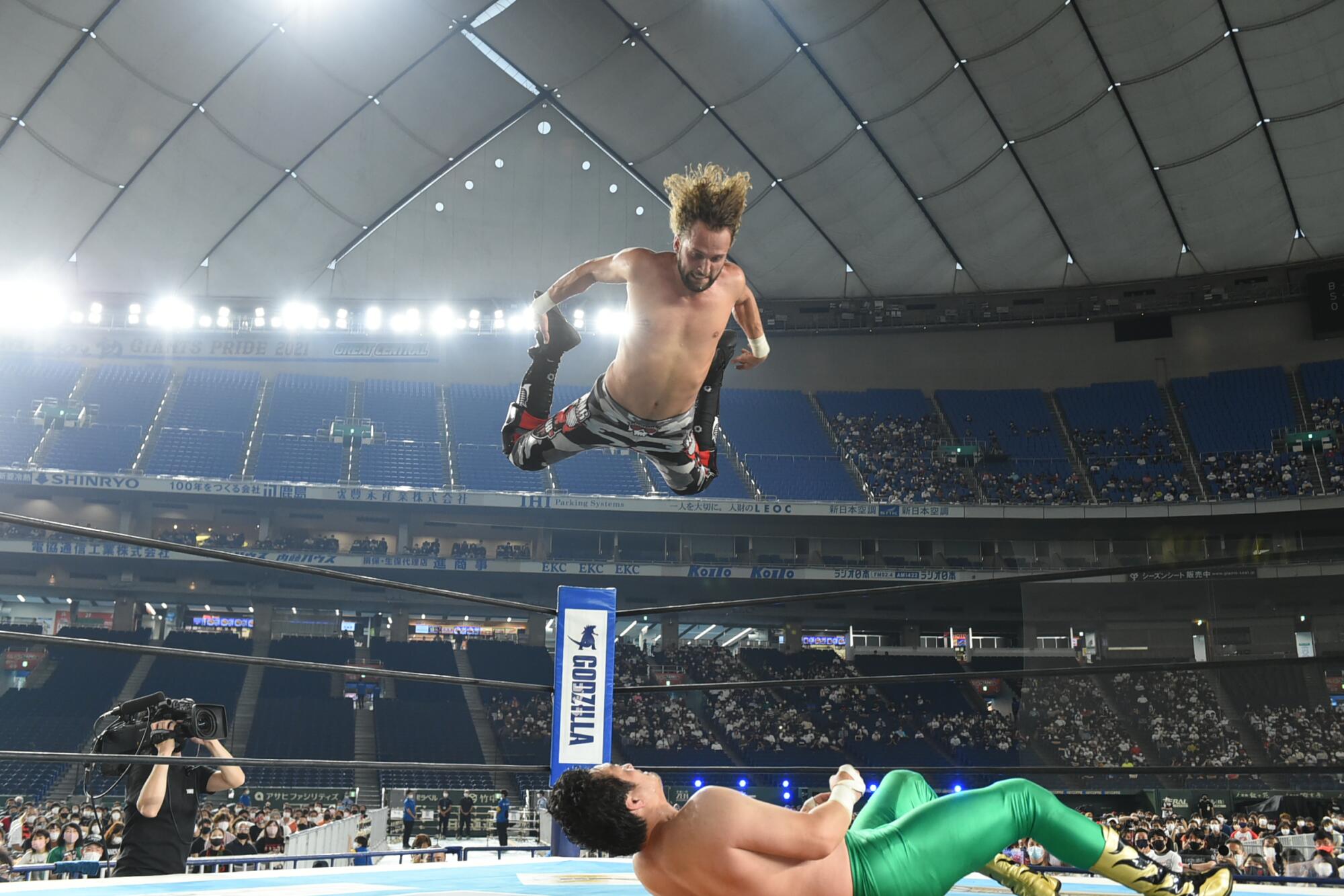"El Phantasmo" leaps midair, high above Ryusuke Taguchi, who lies on the wrestling mat and looks up at him.