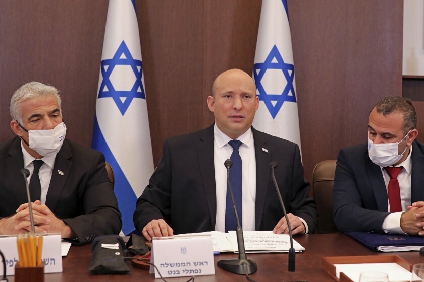 Israeli Prime Minister Naftali Bennett sits at a table with microphones with men flanking him