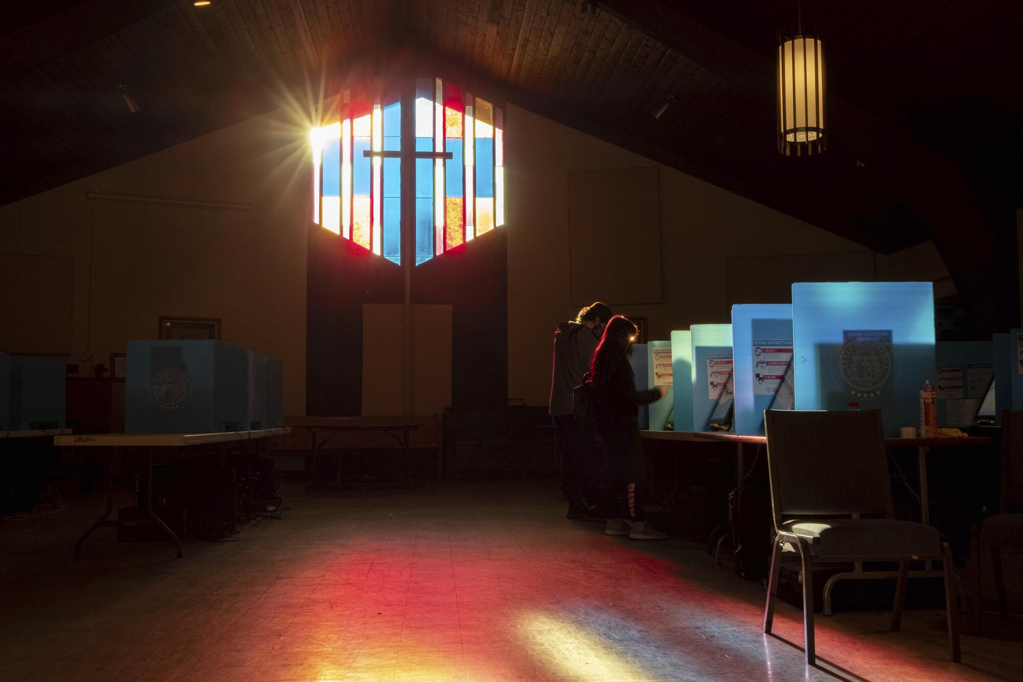 Light glows through a stained-glass window. Two voters in silhouette stand at voting stations.