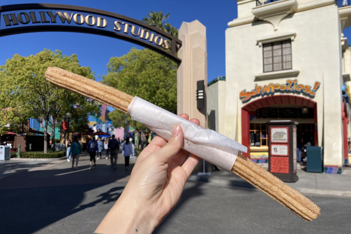 A hand holding a churro at California Adventure in front of a gateway arch that says Hollywood Studios