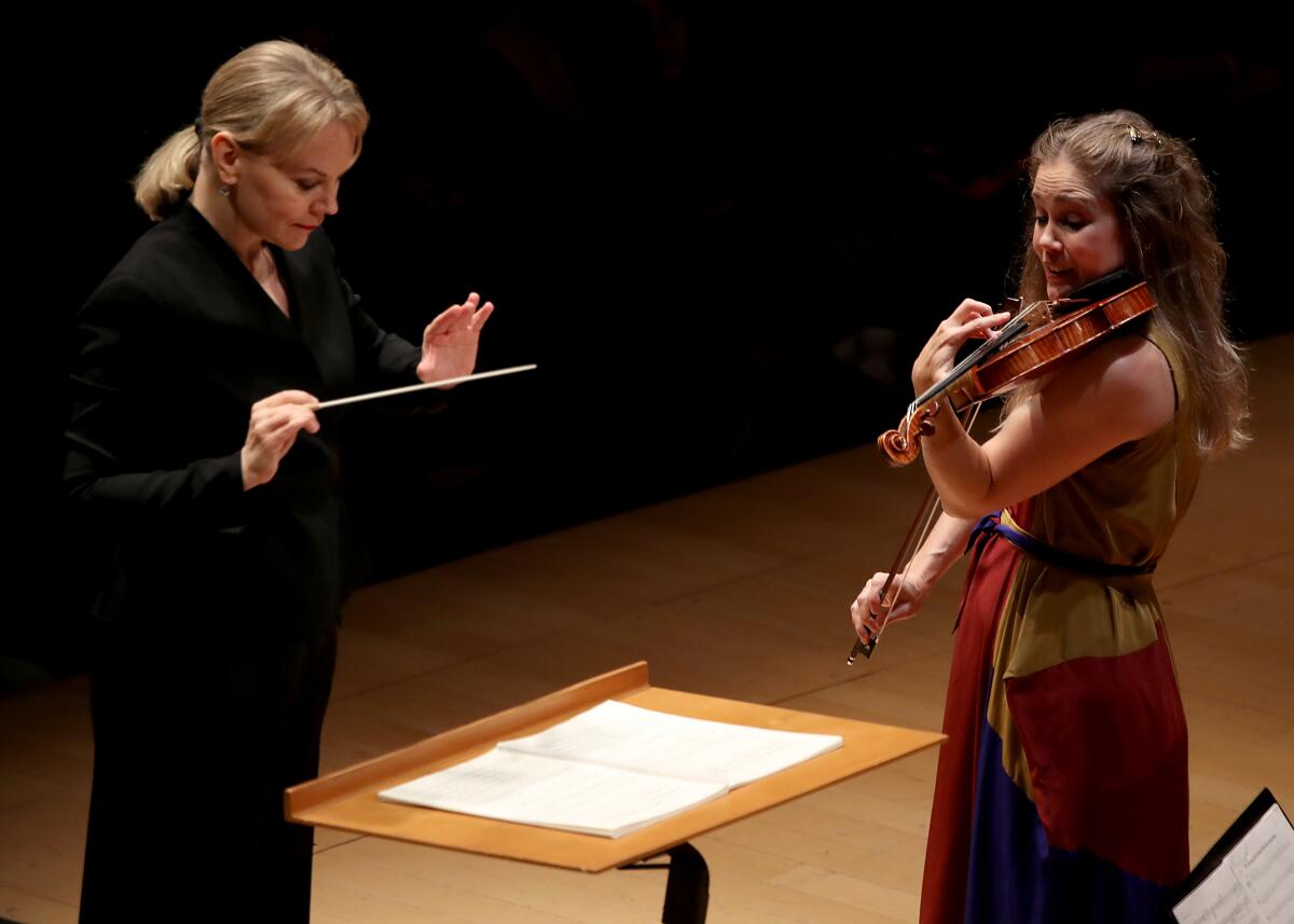 Susanna Malkki is seen in a black suit holding a baton as Leila Josefowicz, in a red dress, plays the violin