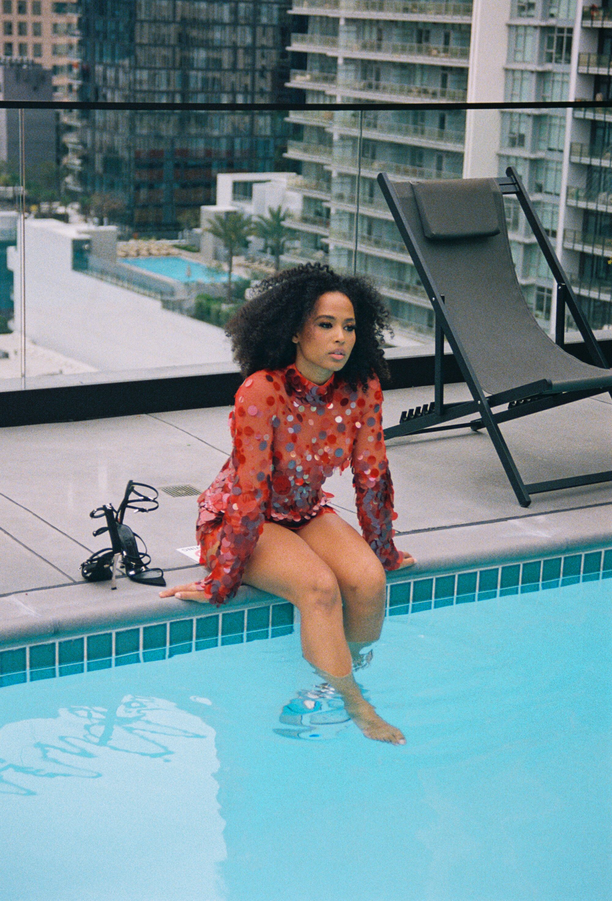 A woman in a red sequined dress sits on the edge of a pool with her feet in the water.