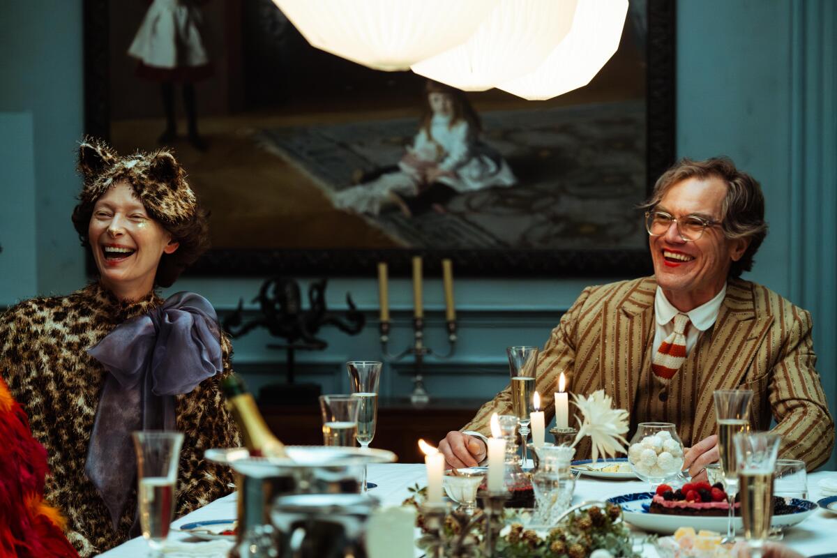 Two smiling people in colorful clothes sit at a dining table.