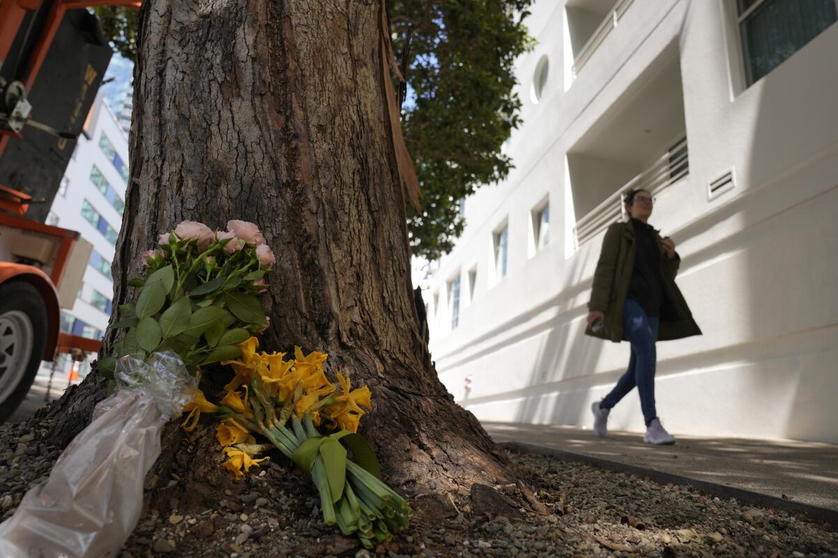 A woman walks past flowers propped against the base of a tree on a city street.