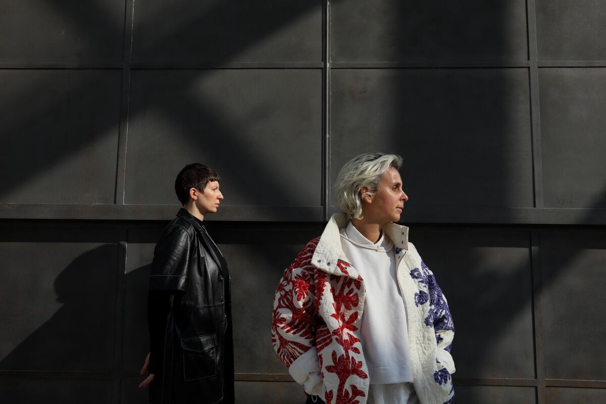 Two people, one in a black leather coat and another in a colorful white coat, stand for a portrait against a dark wall.