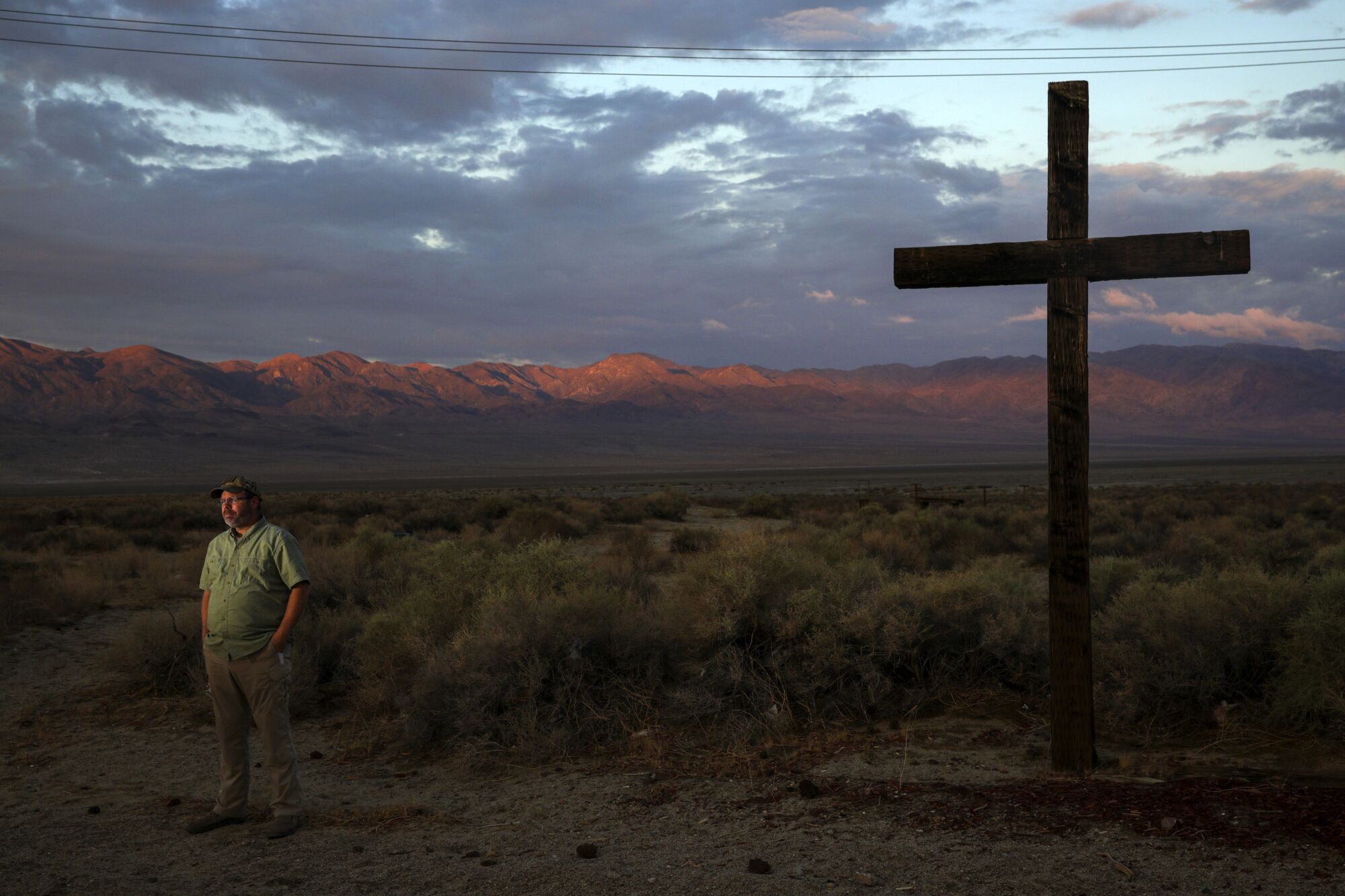 A man stands near a large cross against the backdrop of a desert valley rimmed by mountains