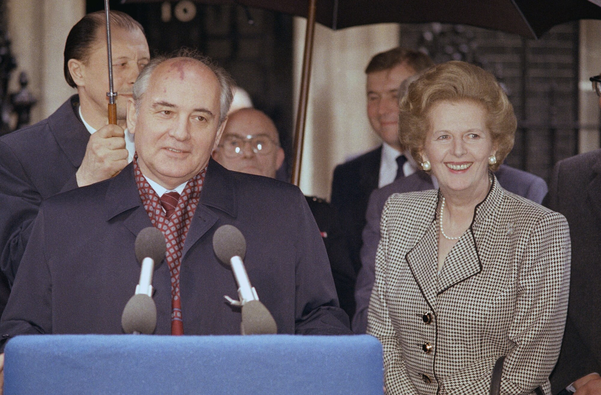 A man in a dark overcoat smiles as he speaks before microphones. With him is a smiling woman, right. 