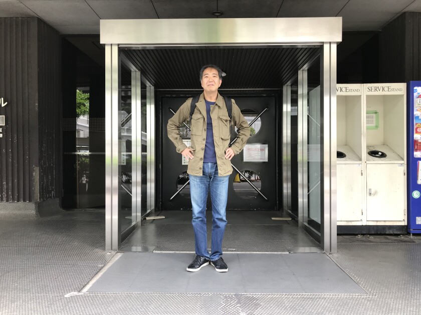 A man in jeans and khaki jacket stands with arms akimbo in front of a city building.