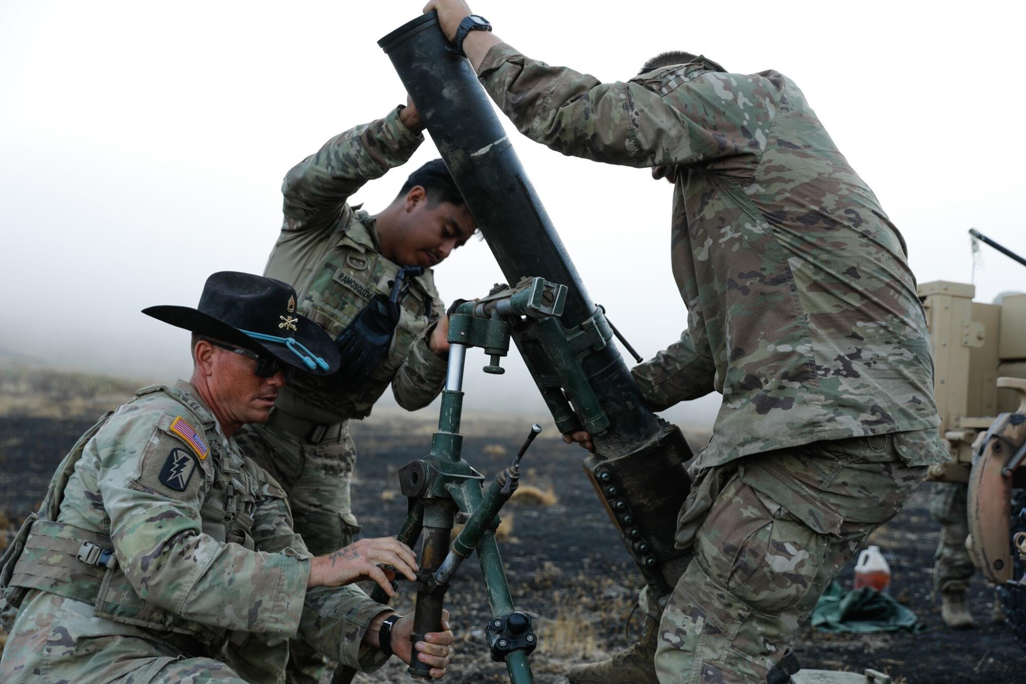 Three people dressed in military uniforms, one of them wearing a black Stetson hat, prepare a mortar to fire.