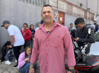 A man with short, dark hair, in a pink long-sleeved shirt, stands near other people gathered outside a building