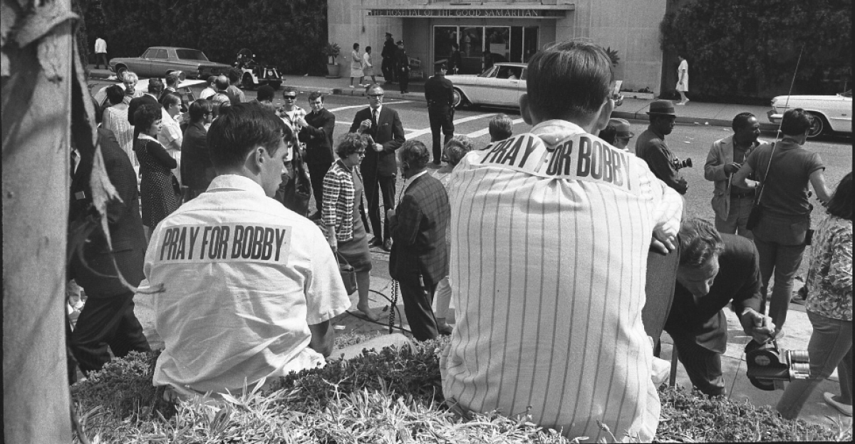 People sit outside Good Samaritan Hospital, two wearing shirts with "Pray for Bobby" signs attached to the backs 