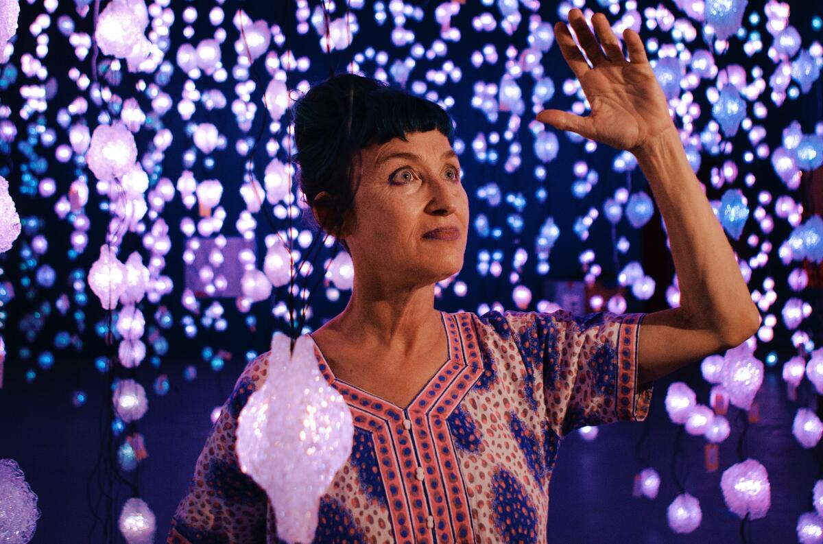 Pipilotti Rist, wearing a patterned tunic, strikes a pose with raised arm in an installation full of blinking LED lights