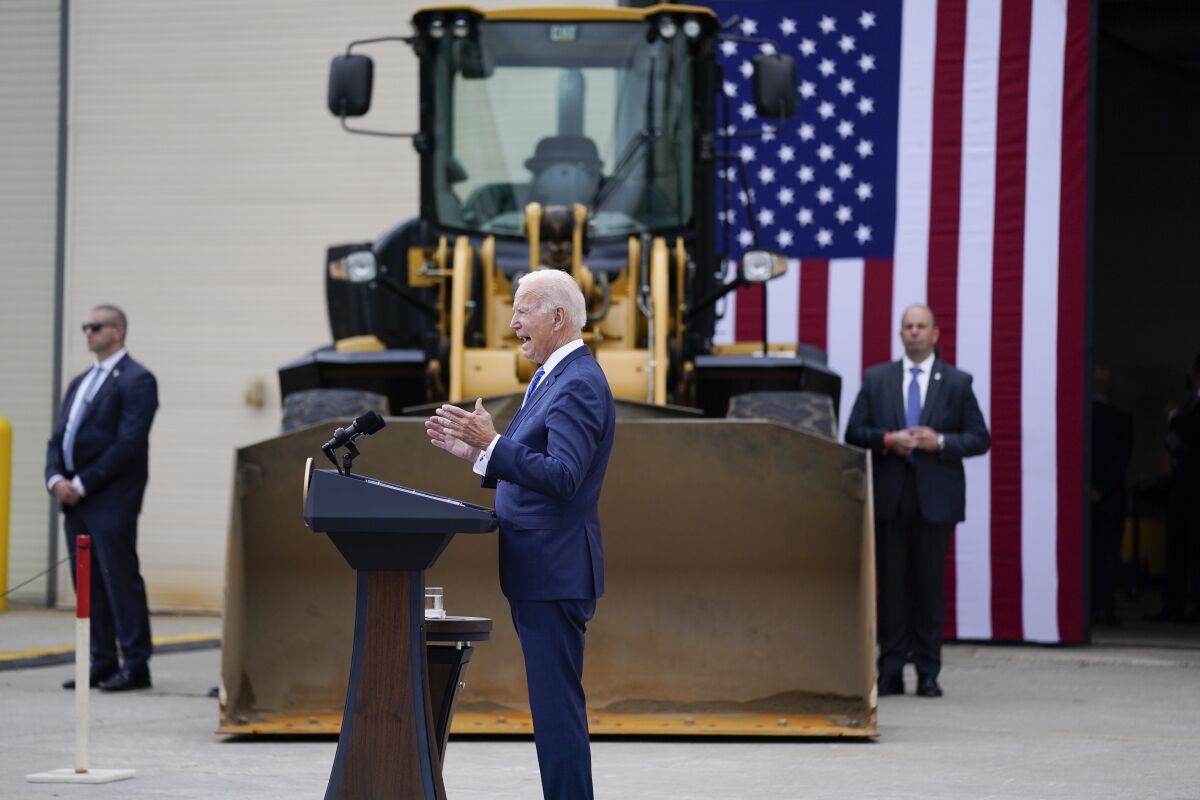 A man in a suit speaks at a lectern in front of heavy equipment flanked by two men, with a U.S. flag in the back
