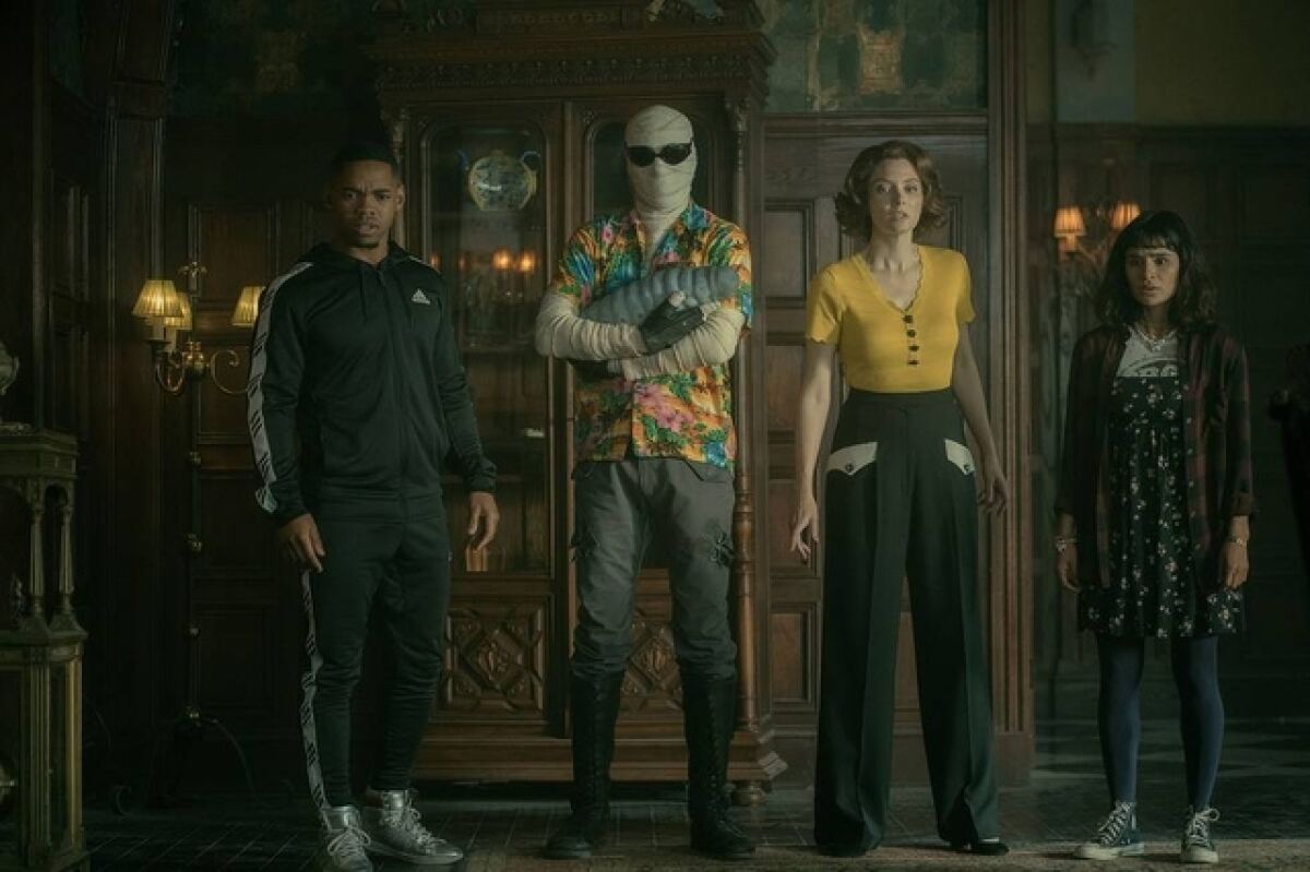 Four people, one in bandages and sunglasses, stand in front of a wood-paneled wall.
