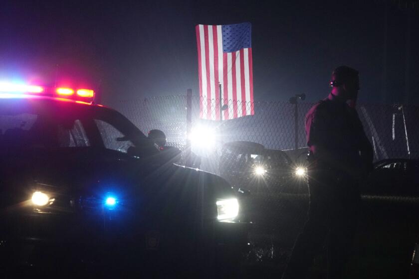 An officer stands at his car early Sunday, July 14, 2024, closing access to the site of the rally where former President Donald Trump was the target of an assassination attempt on Saturday in Butler, Pa.. Behind the officer, a large flag from the rally still flies. (AP Photo/Sue Ogrocki)