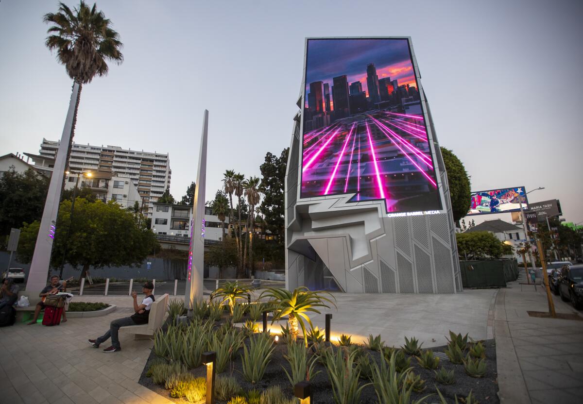 A digital billboard rises from a small parklet and shows images of the Los Angeles skyline on its screen.