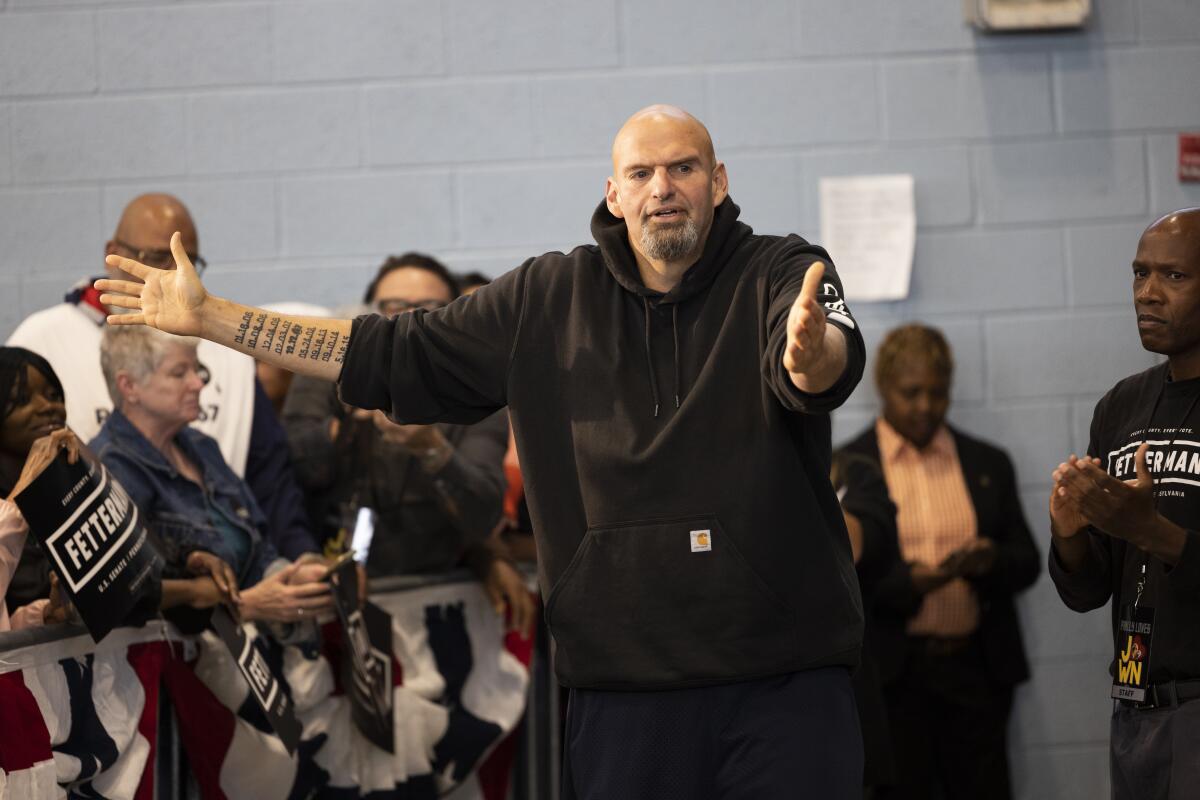 John Fetterman, wearing a hoodie, holds his arms out and gestures to supporters holding his campaign signs. 