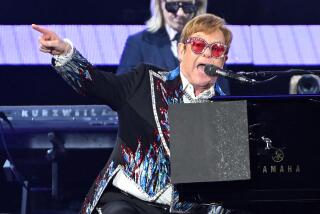 Elton John wears flashy sunglasses and points while playing piano onstage