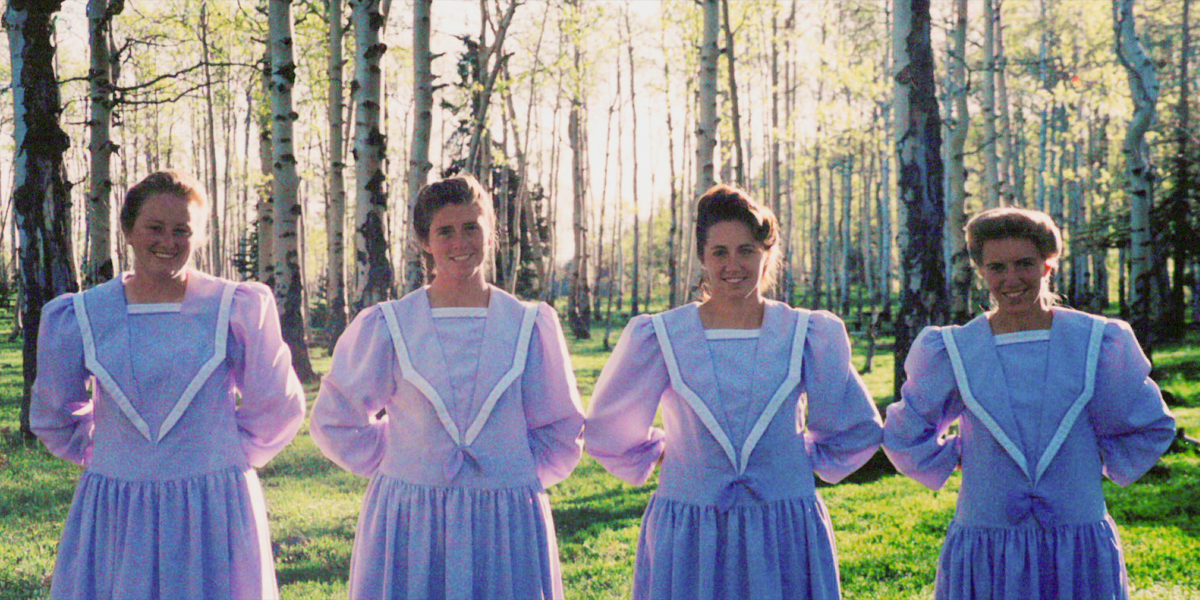 Four women in conservative purple dresses with their hands behind their backs, standing in a wood