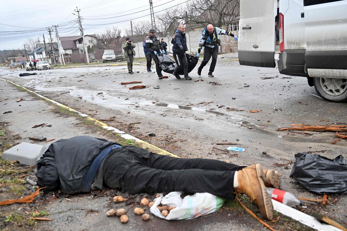 A body lies beside the road as workers carry a body in a bag to a van nearby. 