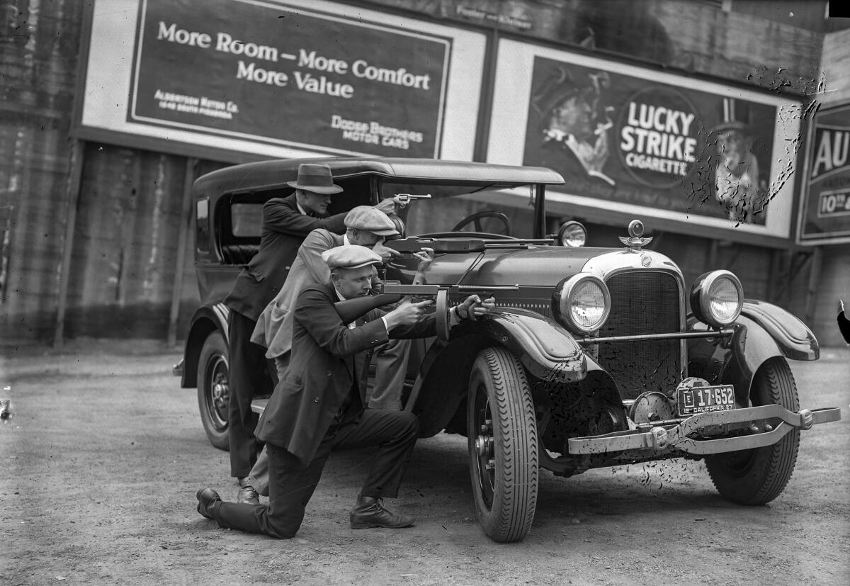Circa 1927: An unpublished photo of three Los Angeles plainclothes policemen crouched behind a car aiming their guns. Handwriting on the negative states "L to R Det. Lt. E.L. Ericksen, Stiles and in front Det. Lt. Oscar Bayer."