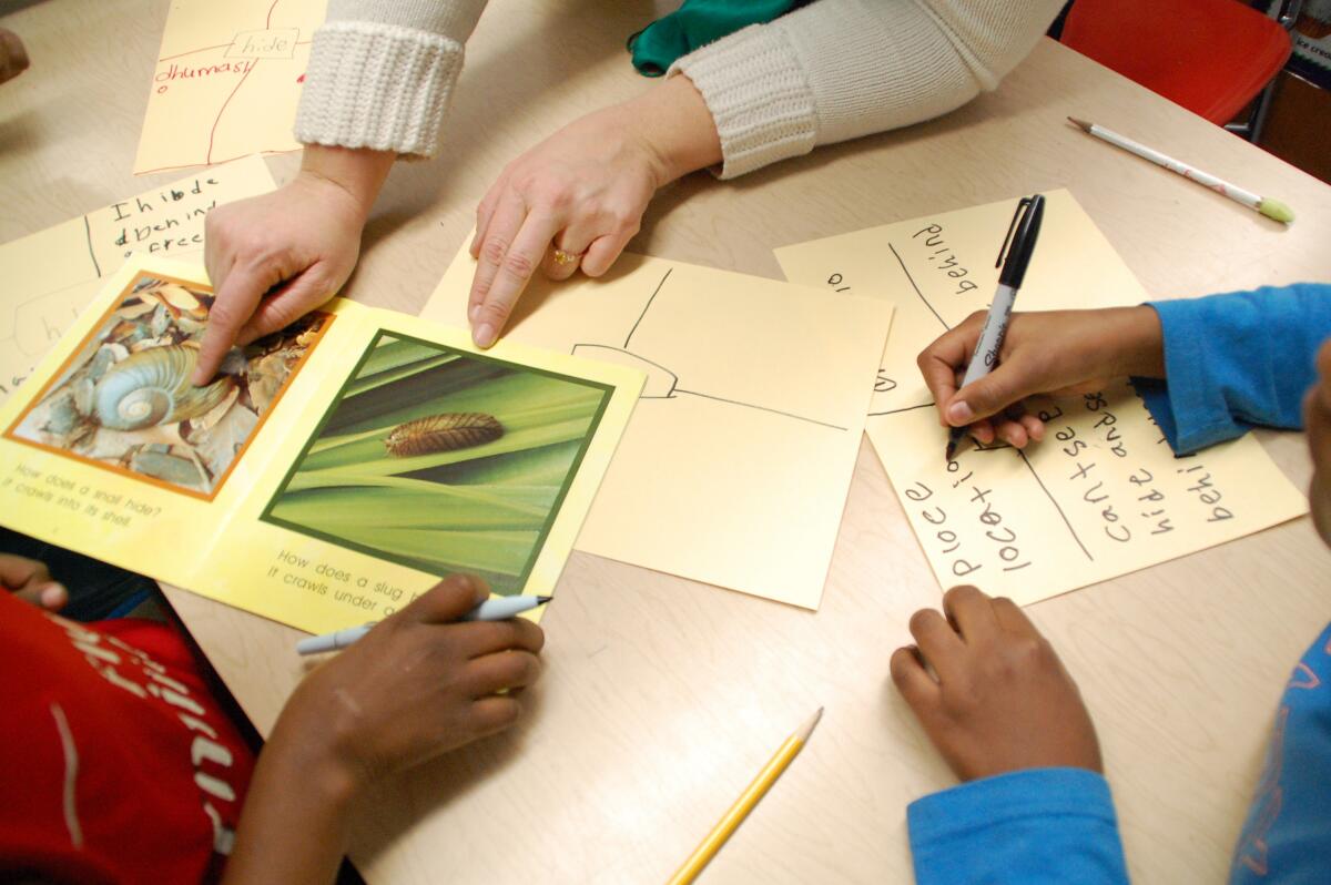 A teacher's hands pointing at pictures on a table, and two students writing or holding pens. 