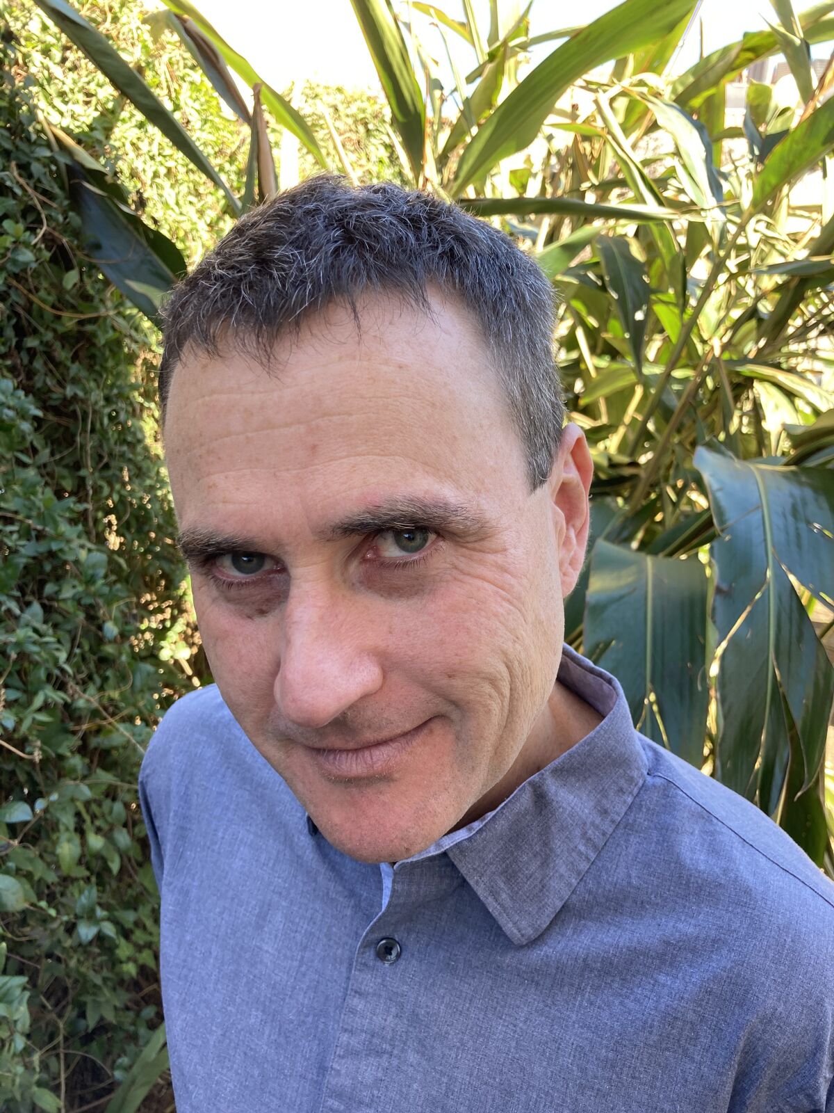 A man in a blue button-down standing among plants looks up at the camera 