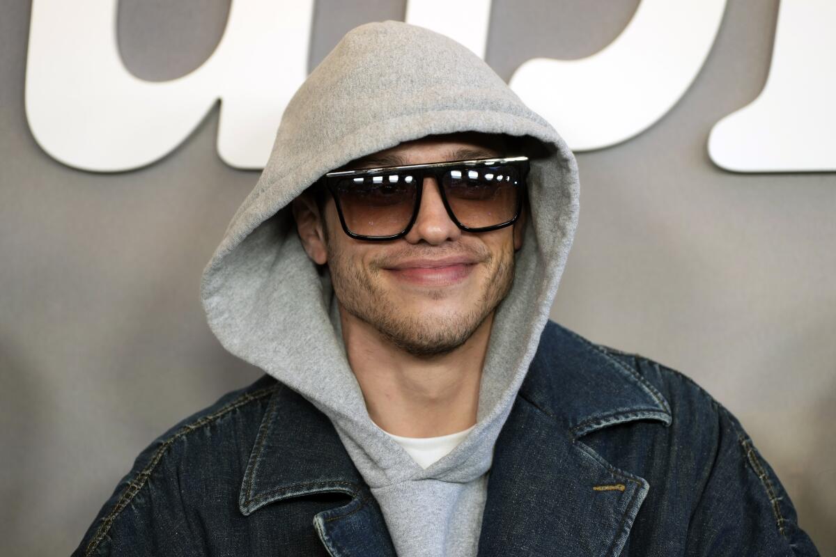 Pete Davidson posing in sunglasses and a blue jean jacket over a gray sweatshirt with the hood up