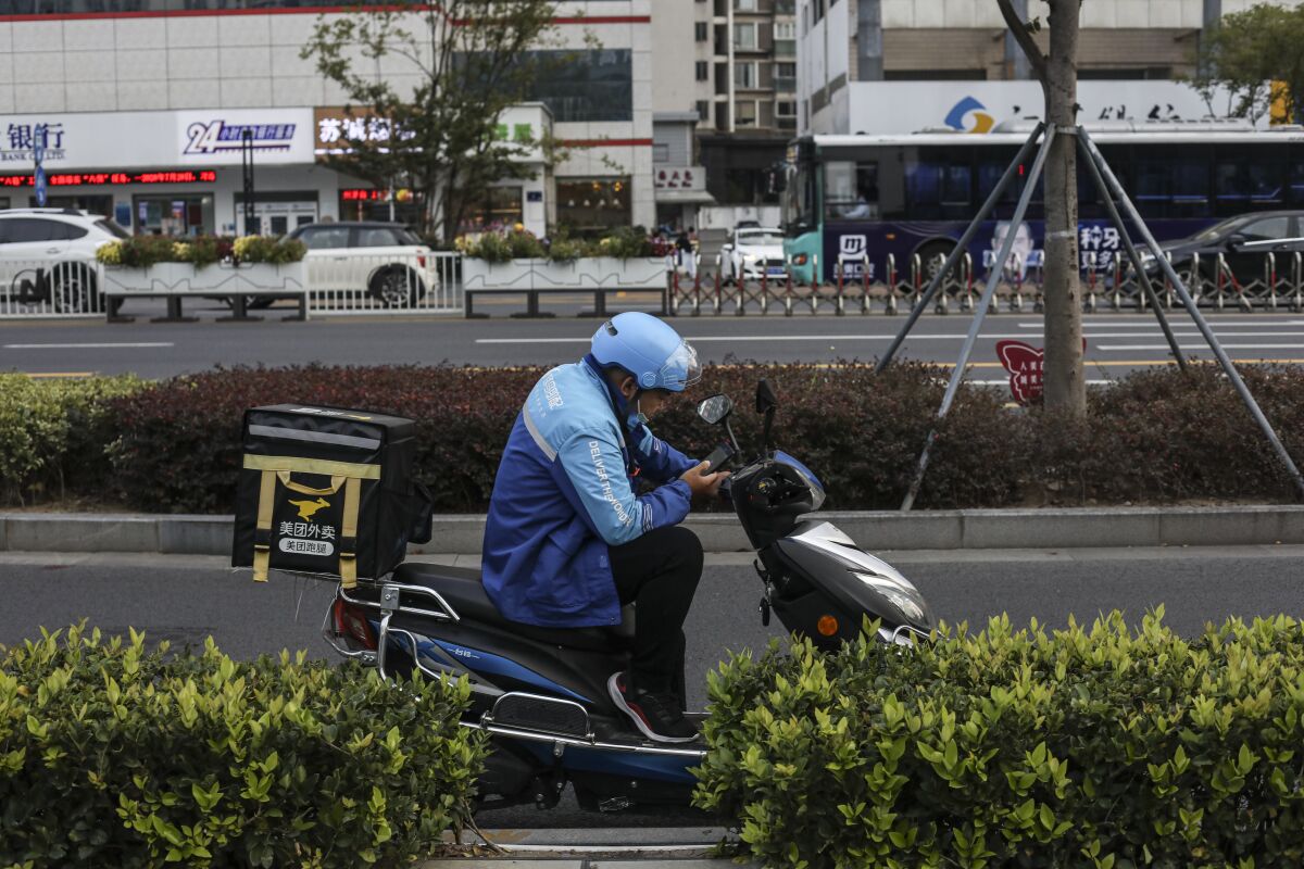 A man on a moped with an insulated delivery bag on the back sits at a curb looking at his phone.
