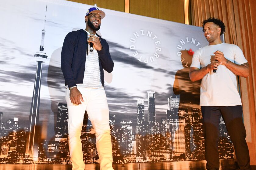 Lebron James holds a microphone and stands next to Maverick Carter on a stage. Both are laughing