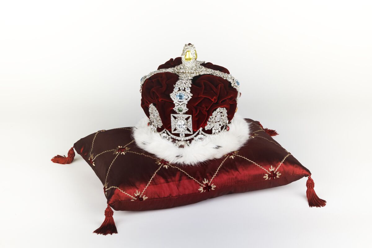 A bejeweled, fur-trimmed crown rests on a red silk pillow that has tassels.