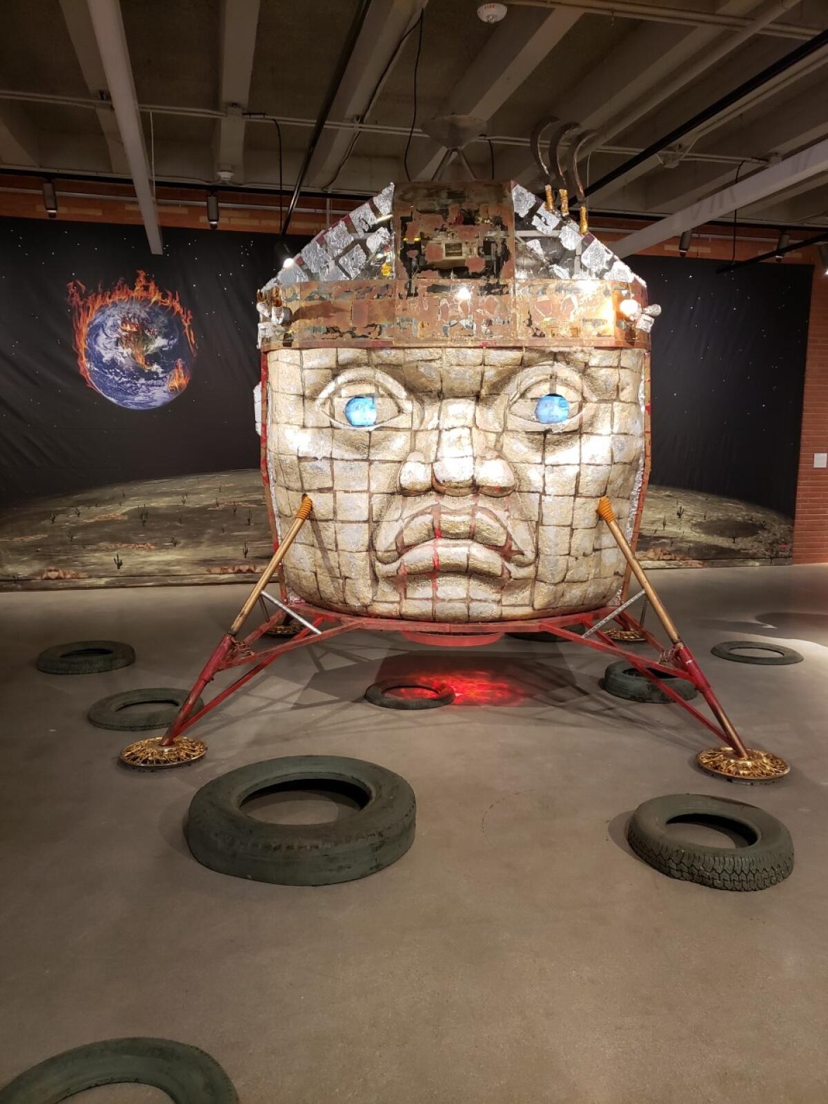 An Olmec head is fused with a lunar landing vehicle with an image of a flaming Earth as backdrop in an art installation.