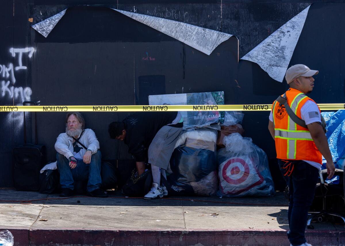 A homeless man with bags sits in the shade against a wall along the sidewalk with yellow police tape in front of him.