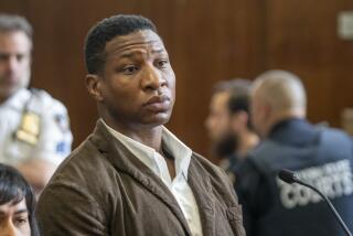Jonathan Majors is standing in a courtroom with a serious face while wearing a brown coat and white shirt.