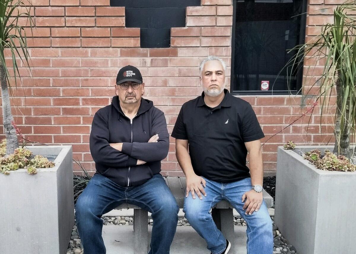 Two men, with the one on the left wearing a cap, sit on a bench outside a building.
