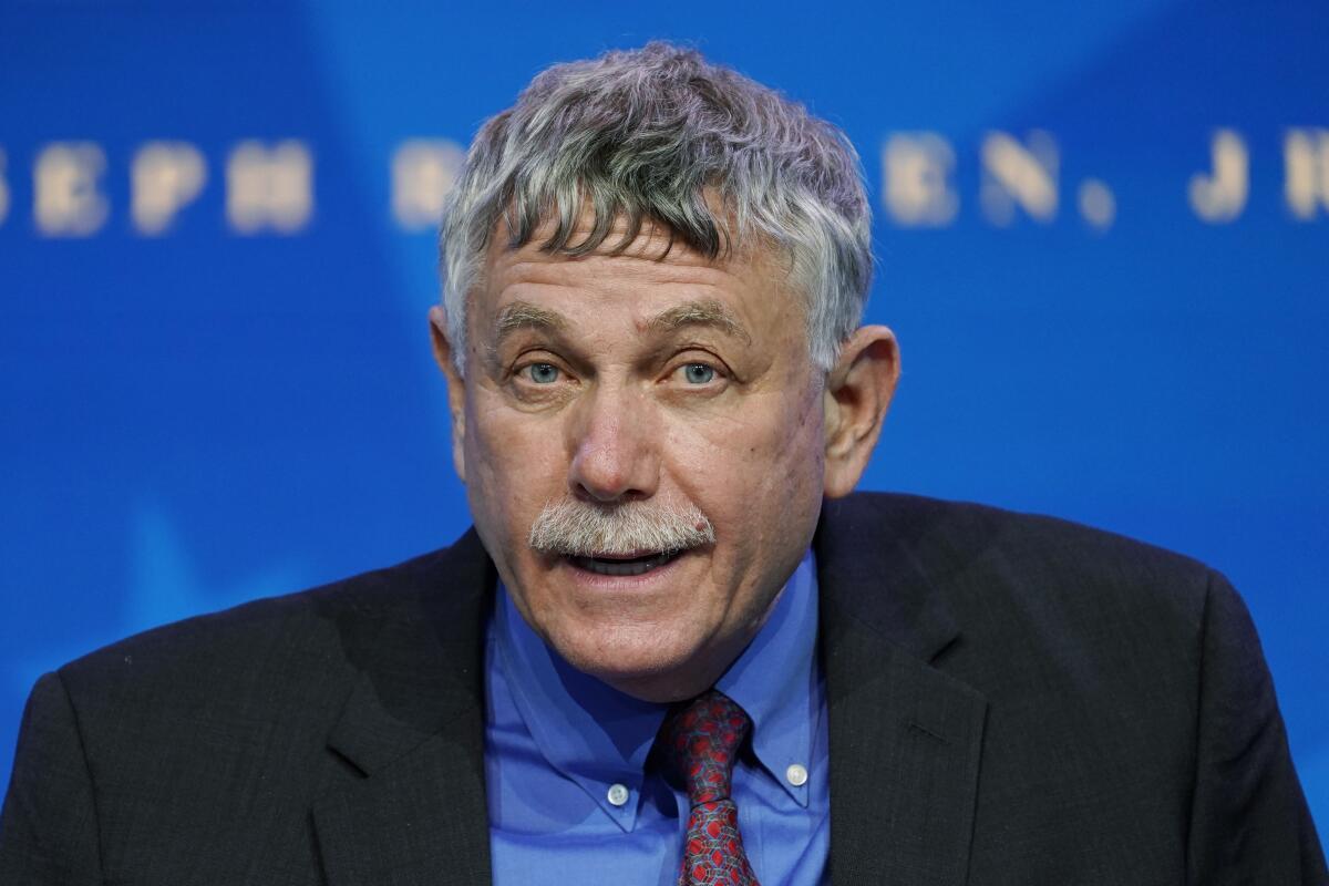 A man with a crooked tie speaks before a blue background.