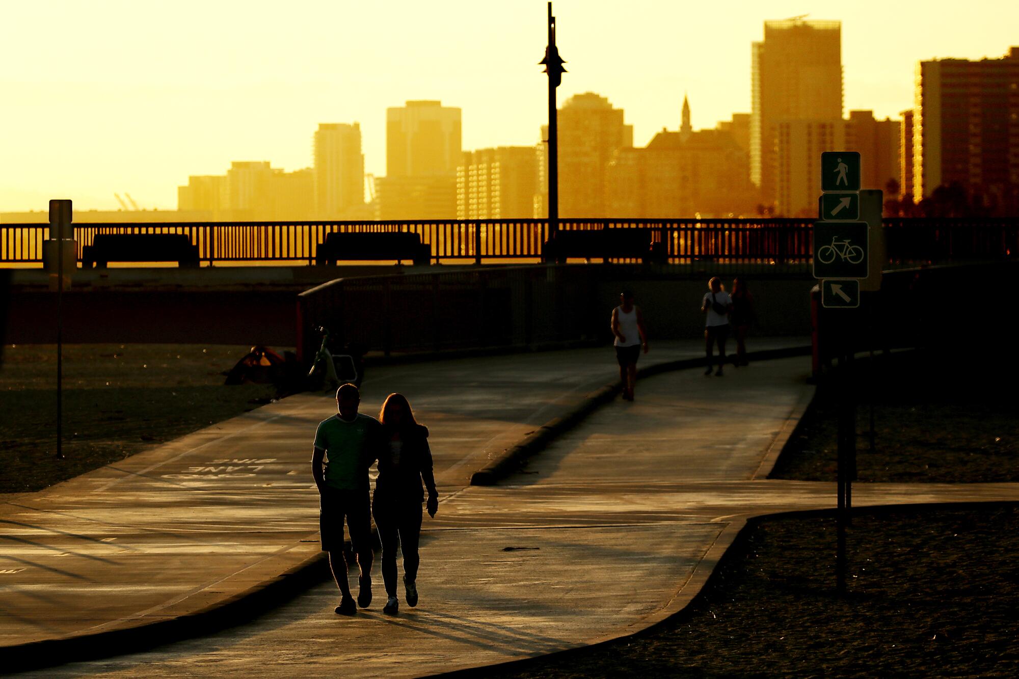 People walk near a pier as the setting sun casts a yellow-orange glow with a city skyline in the background