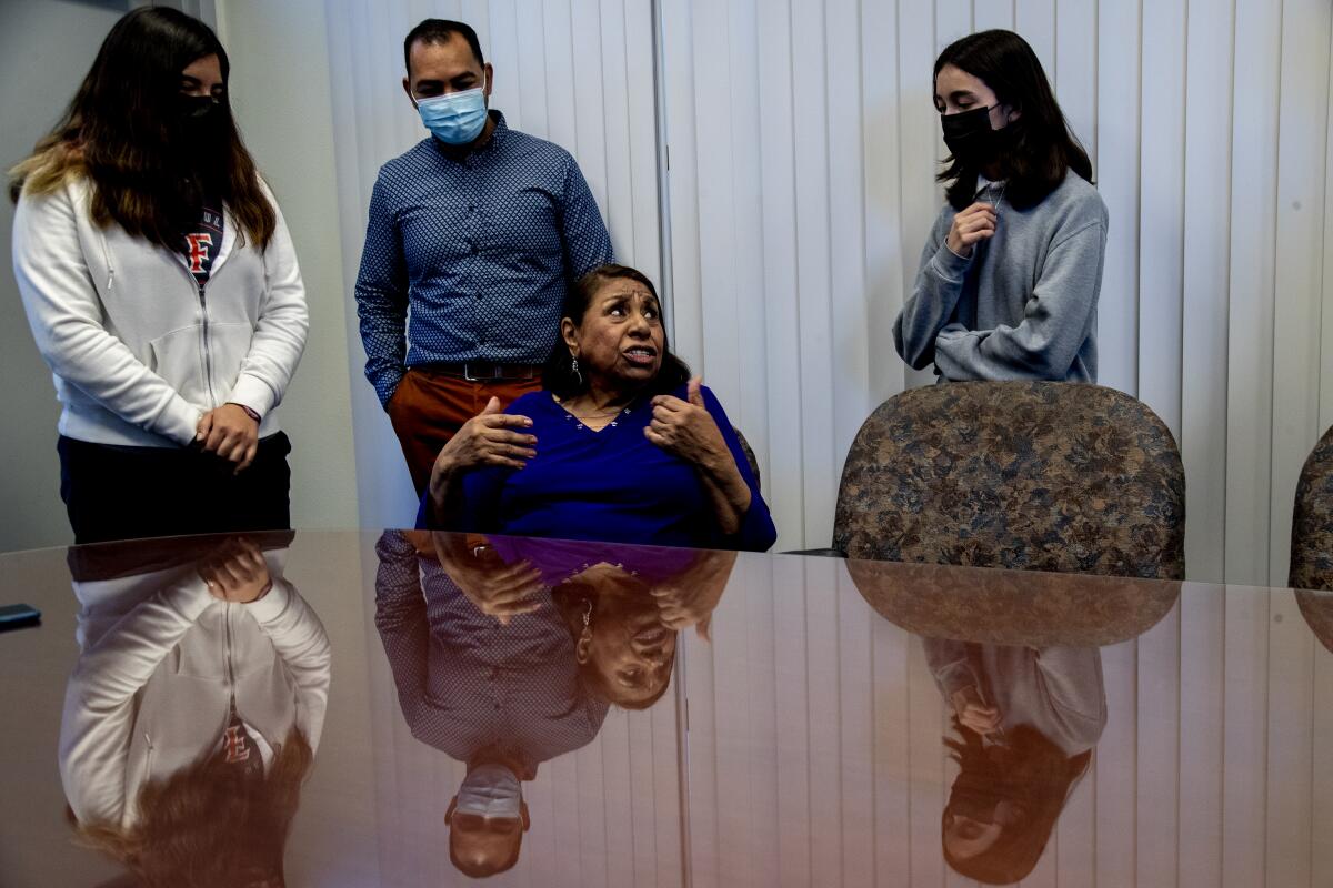 A woman sits at a table with three people standing around her.