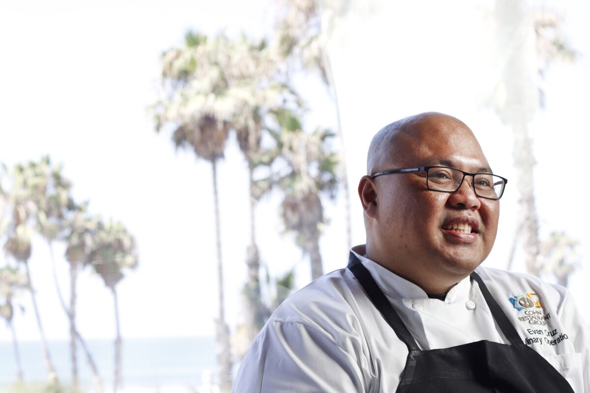A smiling chef sits by a window near the ocean.