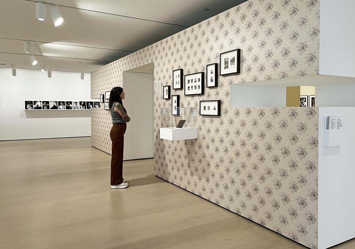 A casually dressed woman examines several small vintage photographs against a gallery wall covered in floral wallpaper.