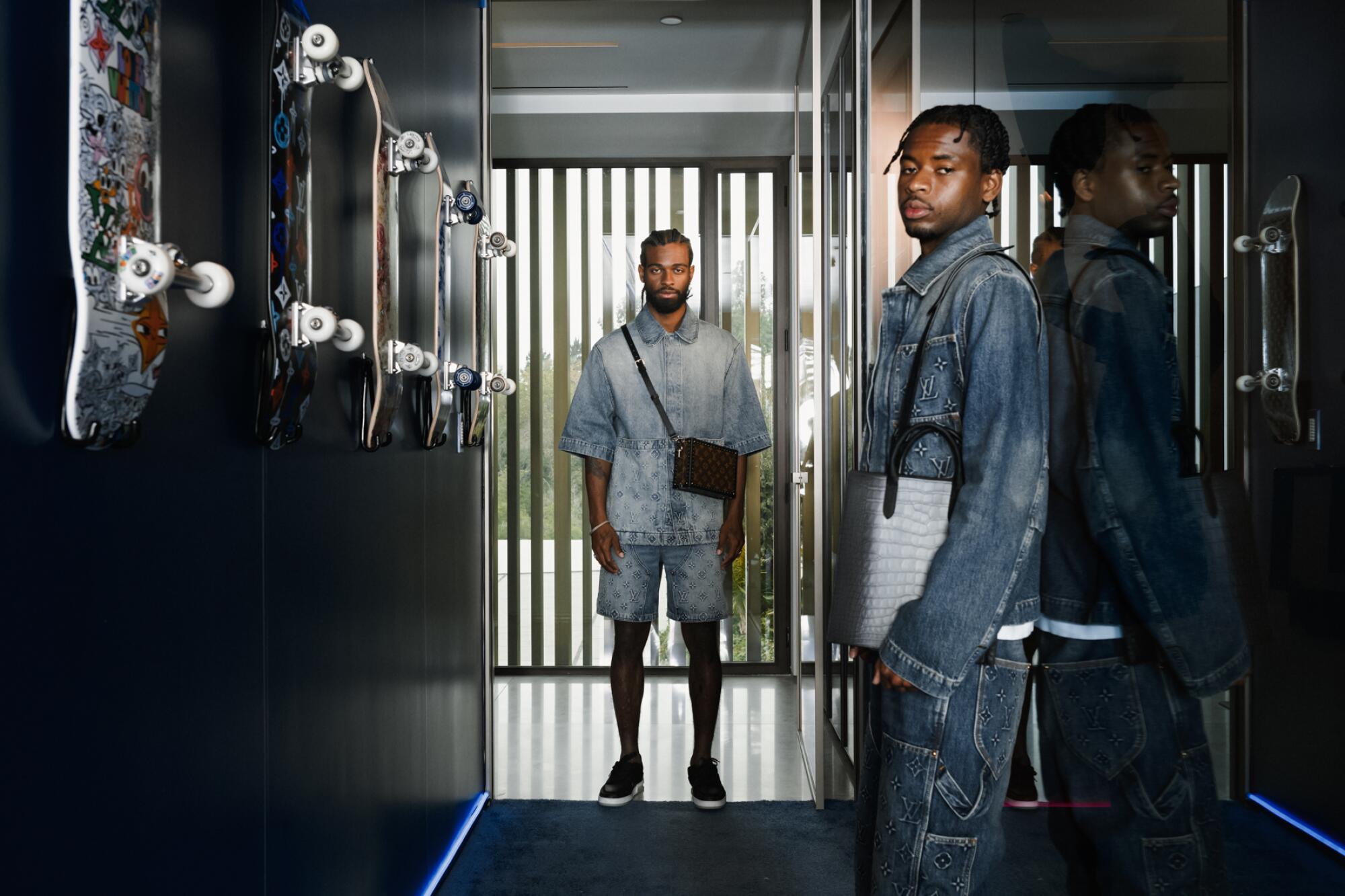 Two men wearing denim pose in a hallway with skateboards on the wall.