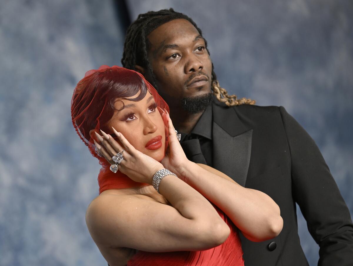 Cardi B with her hands over her ears and wearing a red dress standing by Offset in a black suit