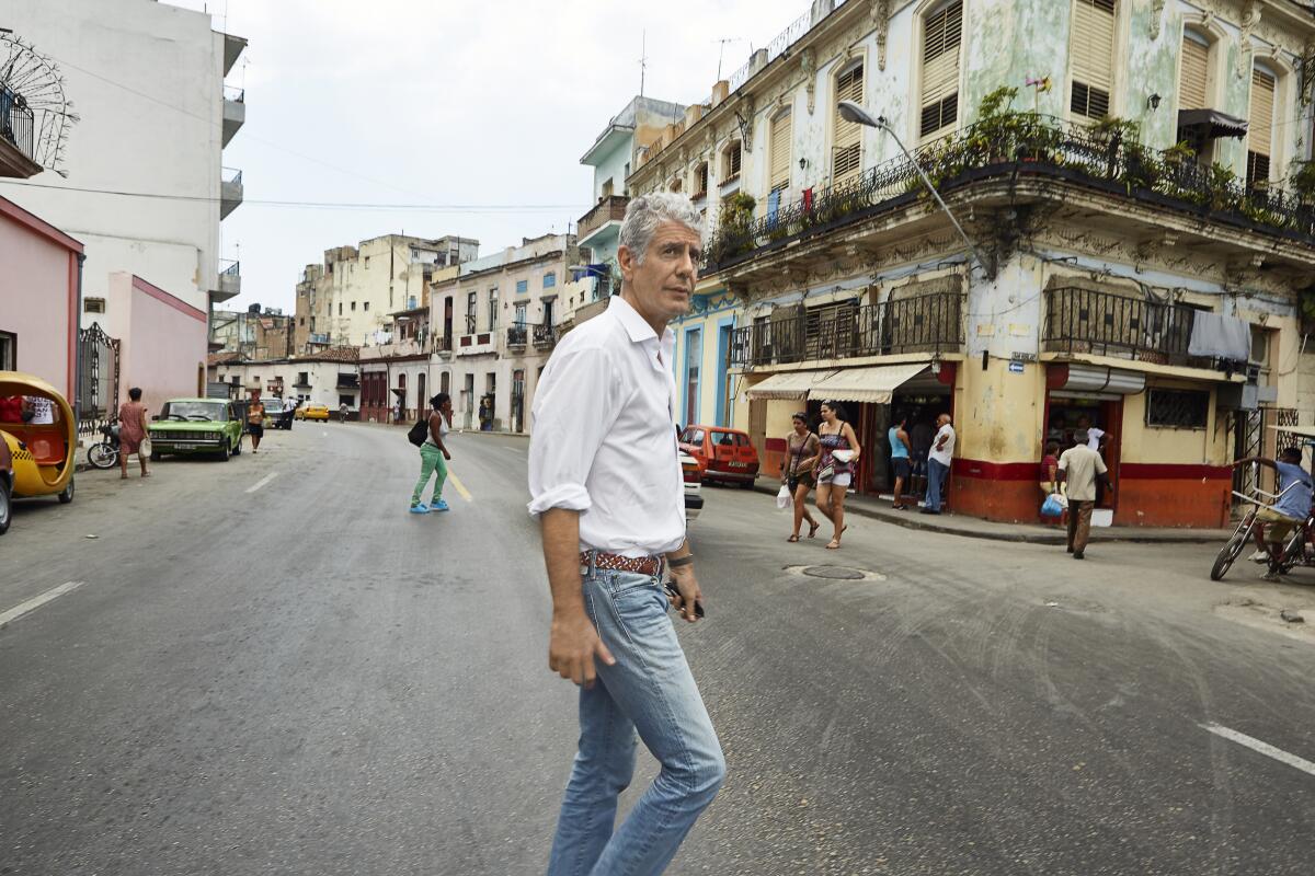 A man wearing a white shirt and jeans stands on a street lined with buildings.