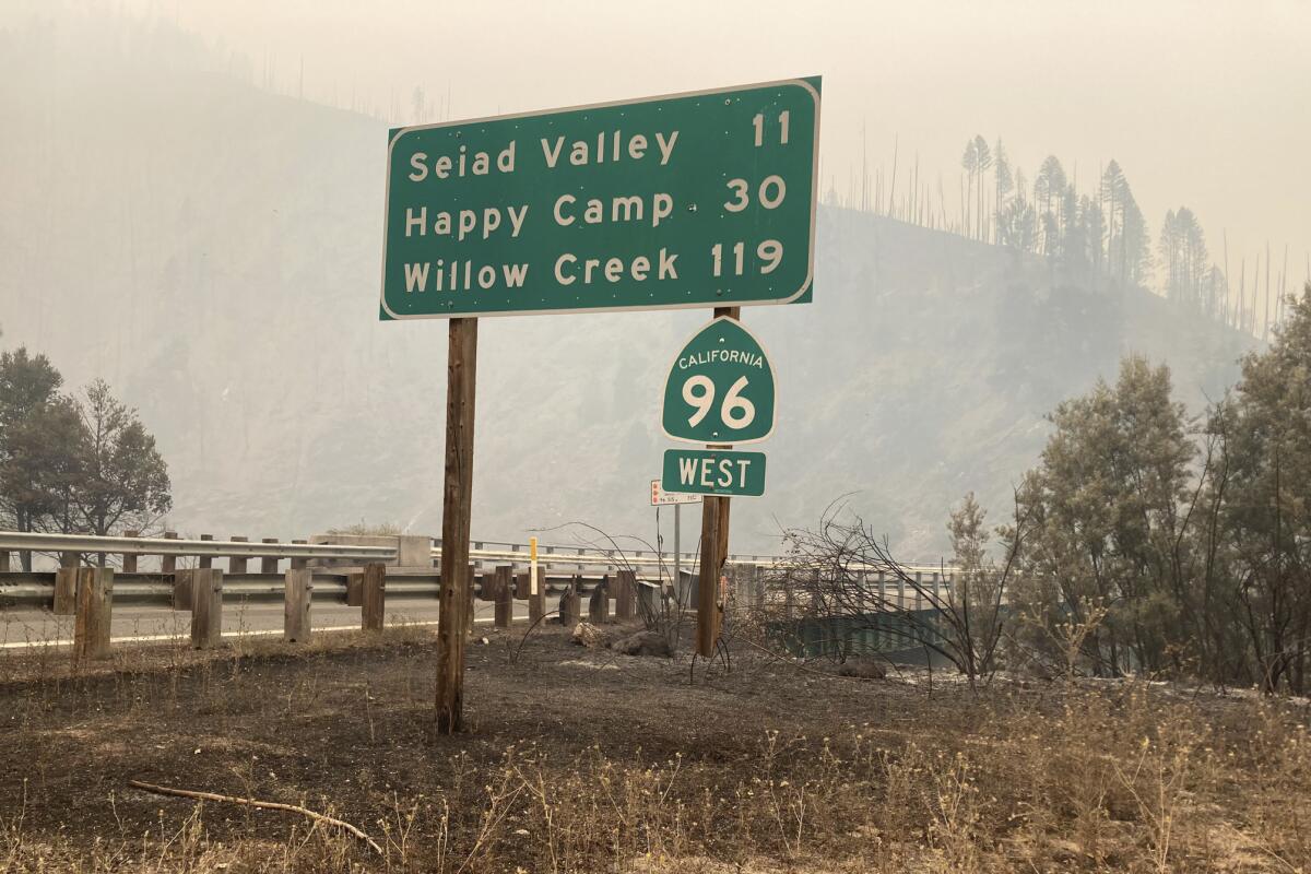 A highway sign on California 96 West shows an exit for Happy Camp in 30 miles, with smoke filling the air behind it