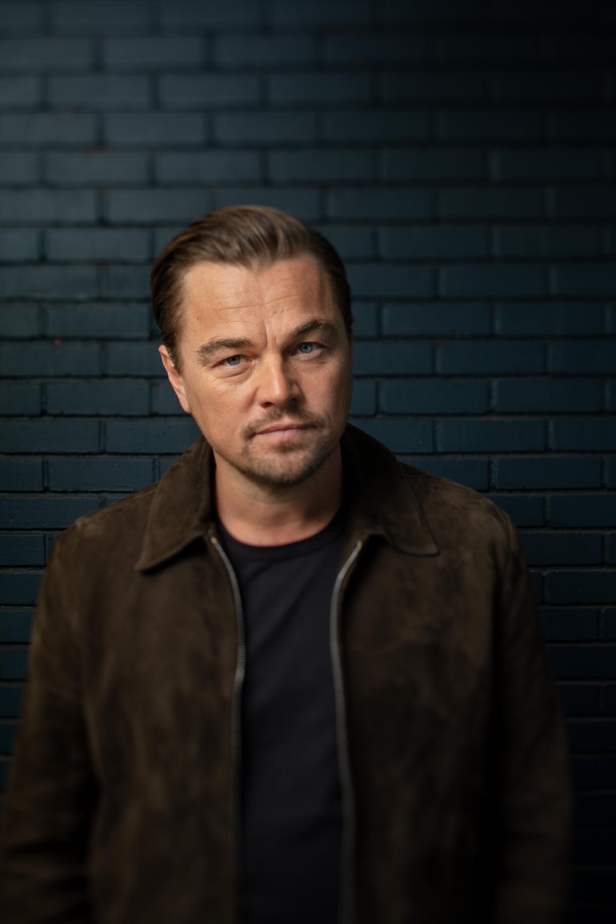 Leonardo DiCaprio stands in front of a dark brick wall for a portrait.