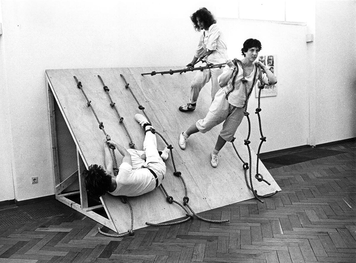 A black and white image shows a group of dancers on a slanted structure, supporting themselves using knotted ropes
