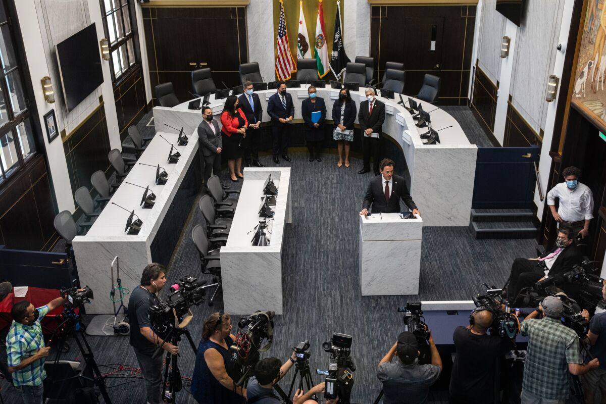 An aerial view of a row of people standing behind a man speaking at a podium, before assembled journalists with cameras.