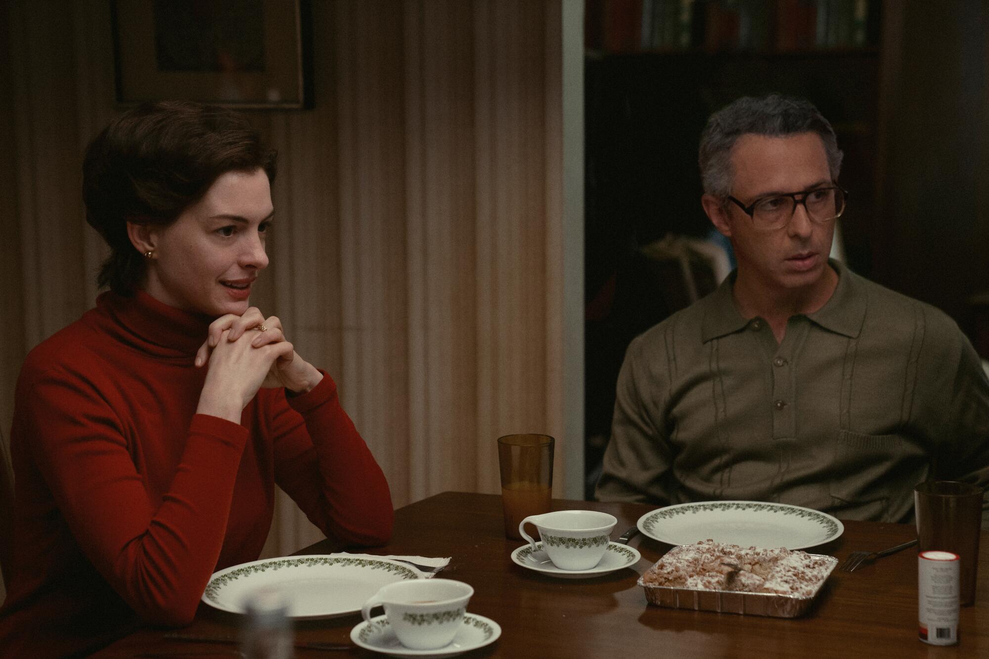 A woman in a red turtleneck, left, and a man in a button-down at a dinner table